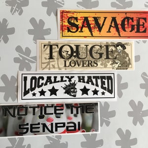 Op de afbeelding: Vier stickers met verschillende ontwerpen. De bovenste sticker is oranje en geel met het woord "SAVAGE" in het zwart. De tweede sticker is beige met zwarte tekst die "TOUGE LOVERS" zegt en een zwart-witte illustratie van een persoon. De derde sticker is wit met zwarte tekst die "LOCALLY HATRED" zegt en drie zwarte sterren. De onderste sticker is wit met zwarte tekst die "NOTICE ME SENPAI" zegt en een afbeelding van een kat.