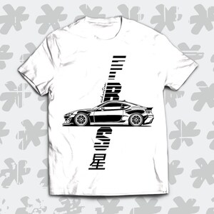 Peut inclure: T-shirt blanc avec une illustration noire et blanche d'une voiture et le texte "EIGHT SIX ZERO" verticalement au centre du t-shirt.