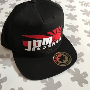 Könnte beinhalten: Schwarze Baseballkappe mit einem roten und weißen Logo, auf dem "JDM" und "日本の国内市場" steht. Das Logo zeigt eine rote aufgehende Sonne. Die Kappe hat einen schwarzen Schirm und einen goldenen Aufkleber mit einem schwarzen Stern an der Seite.