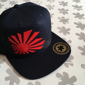 Può includere: Un cappello da baseball blu navy con una grafica di sole nascente rosso sul davanti. Il cappello ha un adesivo dorato con un asterisco nero sul lato.
