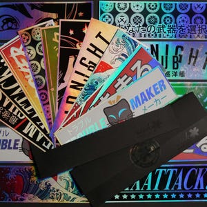 May include: A selection of holographic stickers with Japanese text and graphics. The stickers display various designs, including waves, text that says "NIGHT", "ATTACK!!", and a cat graphic. A black band is also included.