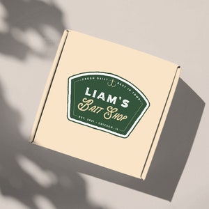 Könnte beinhalten: Ein beigefarbener Karton mit einem grün-weißen Aufkleber, auf dem "Liam's Bait Shop" mit einem Anker-Symbol und dem Text "Fresh Daily - Best in Town - Est. 2021 - Chicago, IL." steht.