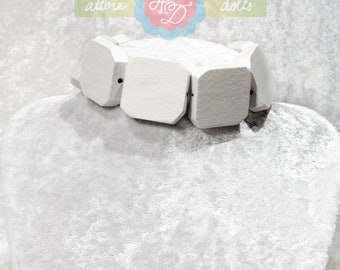 May include: A white, chunky bracelet with geometric, square-shaped wooden beads. The bracelet is displayed on a white, velvet-like surface. The image includes a logo with the words "attire dolls".