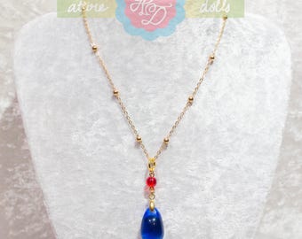 Puede incluir: Collar dorado con colgante azul en forma de lágrima y detalles de cuentas rojas. La cadena fina tiene pequeñas cuentas doradas. El colgante azul oscuro es el punto focal del collar, exhibido sobre un fondo blanco.