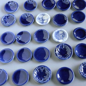 May include: A collection of small, circular ceramic dishes in various shades of deep blue and white. Some pieces feature smooth, glossy surfaces, while others display intricate embossed floral patterns, beaded edges, or mottled white glaze textures.