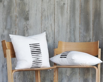 May include: Two wooden chairs with white cushions and black geometric patterns. The chairs are in front of a bookshelf with books and a light pink shelf.