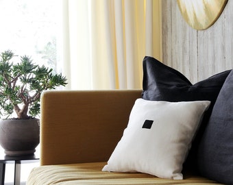 May include: A mustard yellow sofa with three pillows. The sofa has a white pillow with a black square in the center, and two dark gray pillows.