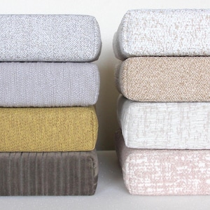 Peut inclure: Quatre coussins empilables de différentes couleurs et textures. Le coussin du haut est gris clair, le deuxième est gris clair, le troisième est jaune moutarde et le dernier est gris foncé. Les coussins sont tous rectangulaires et ont une surface légèrement texturée.