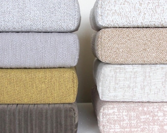 May include: Four stackable cushions in different colors and textures. The top cushion is light gray, the second is a lighter gray, the third is a mustard yellow, and the bottom is a dark gray. The cushions are all rectangular and have a slightly textured surface.