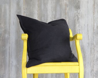 May include: A black linen pillow rests on a bright yellow chair against a gray wooden background. The pillow is square and appears soft. The chair has curved arms and legs, adding a touch of contrast to the scene.