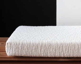 May include: A white, textured cushion sits on a wooden bench. The cushion is made of a soft, woven fabric.