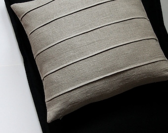 May include: A decorative pillow with a light beige, horizontally-striped design rests on a black fabric. The pillow has a square shape and is made of a textured material. Black ties are visible at the top.