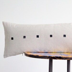May include: A long, rectangular, light gray linen pillow with black square designs. The pillow is resting on a wooden table with a blue and yellow paint design.