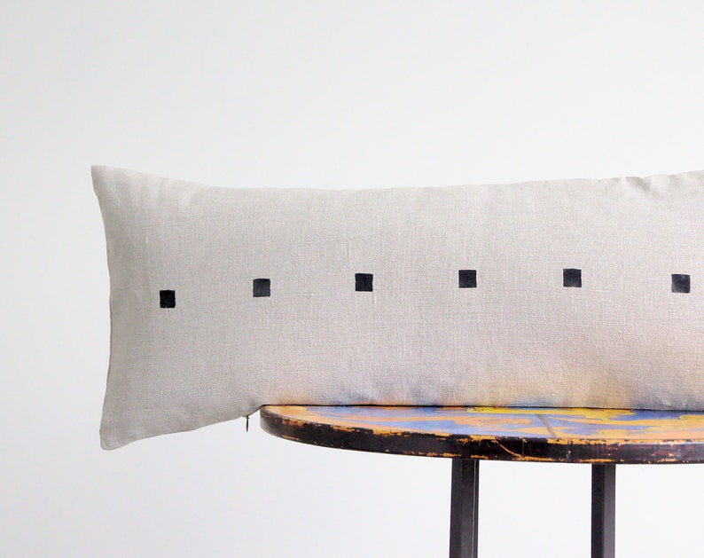 May include: A long, rectangular, light gray linen pillow with black square designs. The pillow is resting on a wooden table with a blue and yellow paint design.