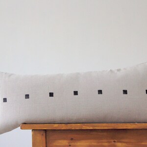 May include: A long, rectangular, light beige linen pillow with a row of black squares across the center.