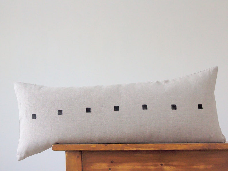 May include: A long, rectangular, light beige linen pillow with a row of black squares across the center.
