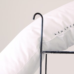 K&ouml;nnte beinhalten: Ein wei&szlig;es Kissen mit dem Text "LINEN SPACE" in Schwarz aufgedruckt. Das Kissen liegt auf einem dunklen Metallrahmen mit einem gebogenen Haken oben. Das Kissen ist einfarbig wei&szlig;.