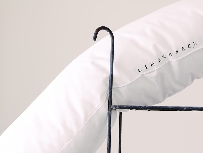 K&ouml;nnte beinhalten: Ein wei&szlig;es Kissen mit dem Text "LINEN SPACE" in Schwarz aufgedruckt. Das Kissen liegt auf einem dunklen Metallrahmen mit einem gebogenen Haken oben. Das Kissen ist einfarbig wei&szlig;.