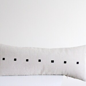 May include: A long, rectangular beige linen pillow with a row of black squares down the center.