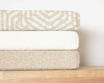 May include: A stack of three rectangular cushions featuring varied textures and colors. The top cushion has a beige and white zebra-like pattern, the middle cushion is solid off-white with a nubby texture, and the bottom cushion is solid beige.