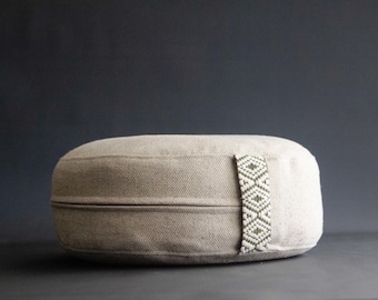May include: A beige, oval-shaped meditation cushion. It features a decorative band with a white and olive green geometric pattern. The cushion is made of a textured fabric and is designed for comfort and support during meditation.