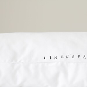 K&ouml;nnte beinhalten: Nahaufnahme eines wei&szlig;en Kopfkissenbezugs mit dem Text "LINENSPACE" in Schwarz gedruckt. Der Kissenbezug besteht aus einem glatten Stoff, wahrscheinlich Leinen. Der Hintergrund ist ein neutrales, hellgraues.