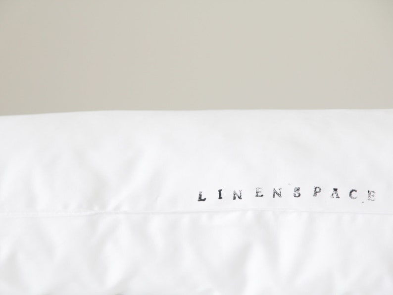 K&ouml;nnte beinhalten: Nahaufnahme eines wei&szlig;en Kopfkissenbezugs mit dem Text "LINENSPACE" in Schwarz gedruckt. Der Kissenbezug besteht aus einem glatten Stoff, wahrscheinlich Leinen. Der Hintergrund ist ein neutrales, hellgraues.