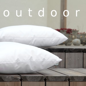 K&ouml;nnte beinhalten: Zwei wei&szlig;e Outdoor-Kissen werden auf einer Holzterrasse gezeigt. Das Wort "outdoor" steht wei&szlig; oben im Bild. Die Kissen sind rechteckig und scheinen aus einem glatten Stoff zu sein.