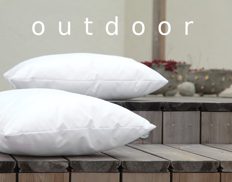 K&ouml;nnte beinhalten: Zwei wei&szlig;e Outdoor-Kissen werden auf einer Holzterrasse gezeigt. Das Wort "outdoor" steht wei&szlig; oben im Bild. Die Kissen sind rechteckig und scheinen aus einem glatten Stoff zu sein.