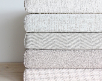 May include: Five different fabric swatches in various shades of beige and white. The swatches are stacked on top of each other, with the top swatch being a plain white fabric and the bottom swatch being a textured beige fabric.