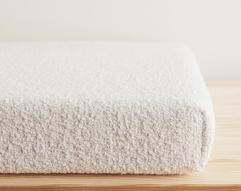 May include: A rectangular cushion or pad covered in a thick, off-white textured bouclé fabric. The material features a dense, nubby, and looped surface texture that creates a soft, voluminous appearance across the entire visible top and side edges.