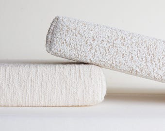 May include: Two rectangular cushions with a textured, off-white fabric surface. One cushion lies flat horizontally, while the other is positioned at an angle, resting one end on top of the flat cushion, showcasing the woven, nubby material.