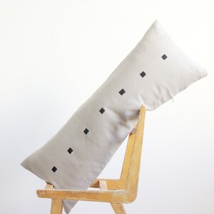 May include: A long, beige linen pillow with black square designs. The pillow is resting on a wooden chair.