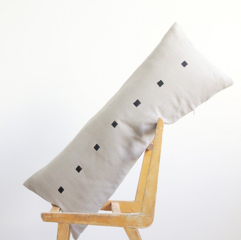 May include: A long, beige linen pillow with black square designs. The pillow is resting on a wooden chair.