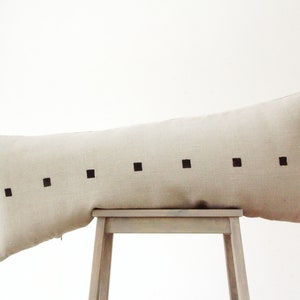 May include: A long, beige linen pillow with a row of black squares down the center.