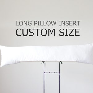 K&ouml;nnte beinhalten: Ein langes, wei&szlig;es Kissen, das auf einem schwarzen Metallst&auml;nder pr&auml;sentiert wird. Das Kissen ist rechteckig und scheint mit einem weichen Material gef&uuml;llt zu sein. &Uuml;ber dem Kissen steht der Text "LONG PILLOW INSERT CUSTOM SIZE."