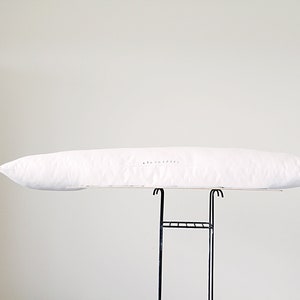 K&ouml;nnte beinhalten: Ein langes, wei&szlig;es Kissen mit dem gestickten Text "a&e lofted" an der Seite. Das Kissen wird auf einem schwarzen Metallgestell vor einem schlichten wei&szlig;en Hintergrund pr&auml;sentiert. Das Kissen ist etwa 91 cm lang.