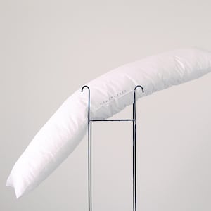 May include: A long, white body pillow rests on a black metal stand. The pillow is angled, showcasing its shape and texture. The pillow has the word "SQUAREFACT" printed on it. The background is a plain, light color.