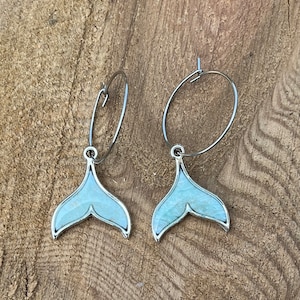 May include: A pair of silver hoop earrings with a mermaid tail design. The tail pendants are light blue with a silver outline, set against a wooden background. The earrings are a unique accessory.