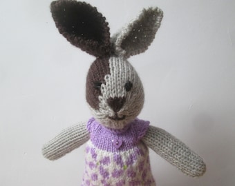 May include: A knitted gray and brown bunny wearing a purple and white floral dress with a purple button.