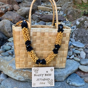 May include: A woven straw tote bag with a black and gold lei. The bag has a wooden tag that says "Busy Bee Studio 2014".