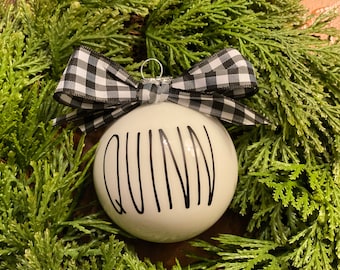 May include: A round white ornament featuring the name QUINN written in black capital letters. The ornament is topped with a black and white checkered fabric bow and a silver metal hanging loop, creating a decorative piece for seasonal display.