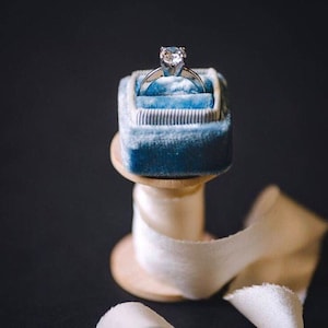 May include: A diamond engagement ring rests in a teal velvet ring box. The ring box is displayed on a wooden spool with a white ribbon. The background is a dark, solid color.