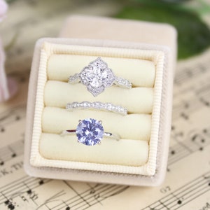 May include: A white velvet ring box with three rings inside. The top ring is a silver ring with a large, clear, round stone surrounded by smaller stones. The middle ring is a silver band with a delicate design. The bottom ring is a silver ring with a large, purple, round stone.