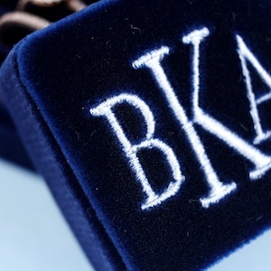 Puede incluir: Una caja de terciopelo azul marino con las iniciales "BKA" bordadas en blanco.