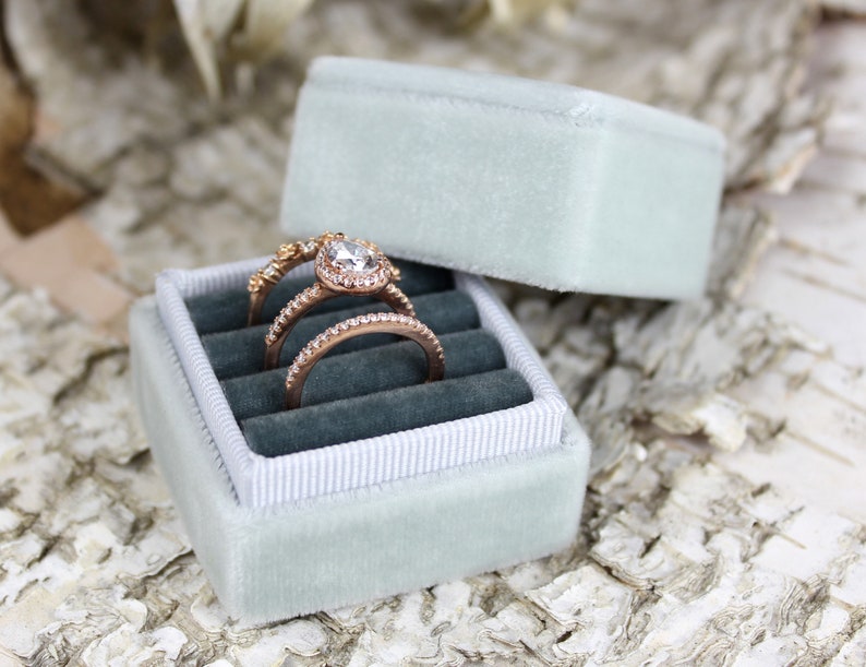 Puede incluir: Una caja de anillo de terciopelo azul claro con un forro de sat&eacute;n blanco. Tres anillos de oro est&aacute;n dentro de la caja, incluyendo un anillo de compromiso de forma de pera con un engaste halo.