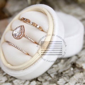 Puede incluir: Un conjunto de tres anillos de oro rosa presentados en una caja de anillos de terciopelo blanco. El conjunto incluye un anillo de compromiso con halo en forma de pera y dos alianzas de boda. Los anillos están engastados con piedras preciosas claras. La caja está abierta, mostrando los anillos.