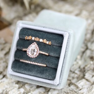 Puede incluir: Tres anillos de oro con diamantes en una caja de anillo de terciopelo azul. El anillo central presenta un diamante en forma de pera.