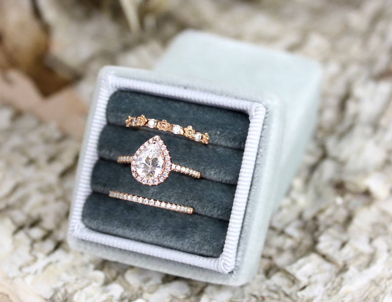 Puede incluir: Tres anillos de oro con diamantes en una caja de anillo de terciopelo azul. El anillo central presenta un diamante en forma de pera.