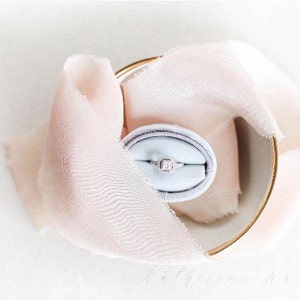 May include: A delicate engagement ring with a square-shaped diamond set in a silver band, displayed in a light blue velvet ring box. The box is nestled in a soft, pink fabric.